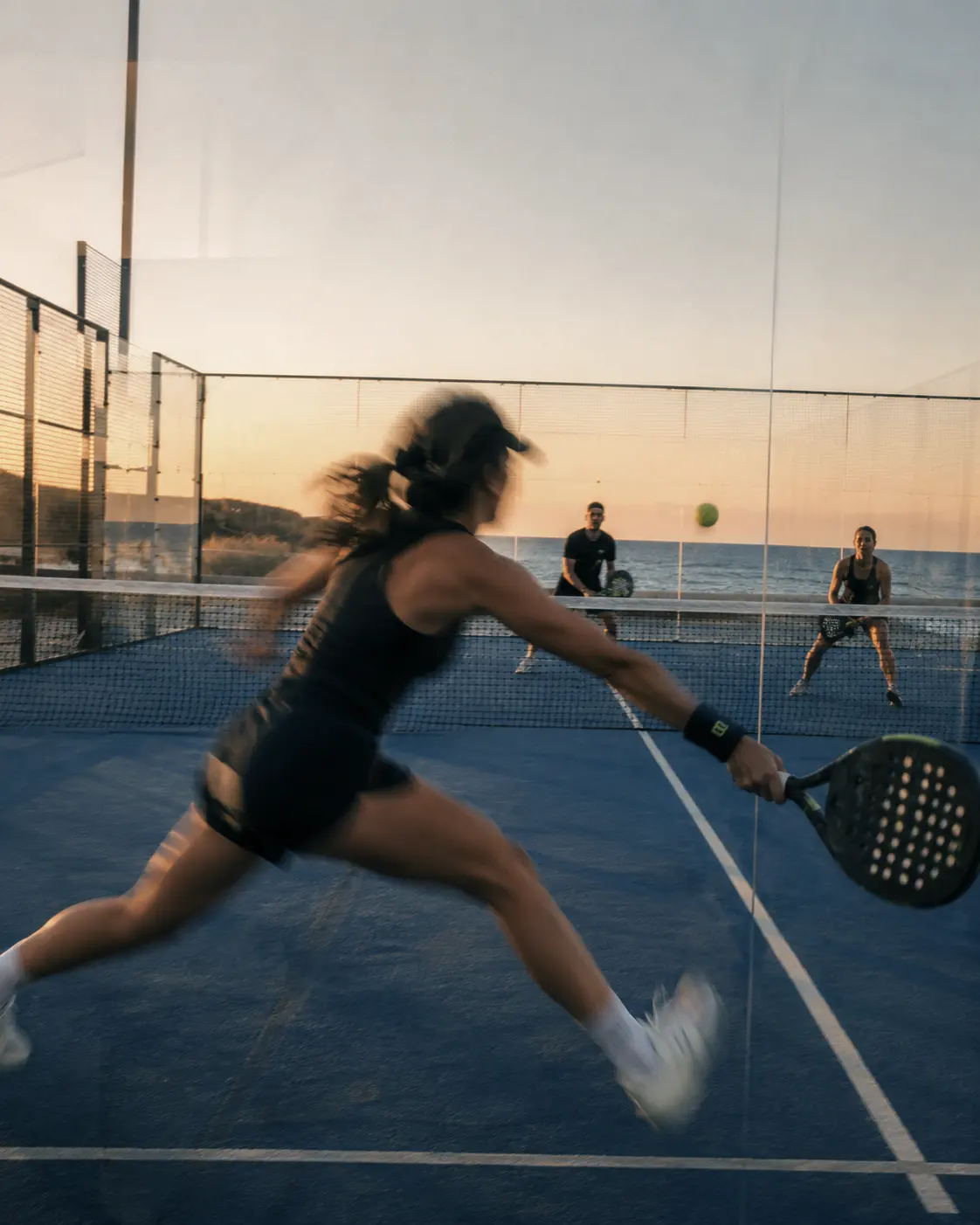 Court No. 01 at last light, a player reaching for a low ball with the sea beyond the glass