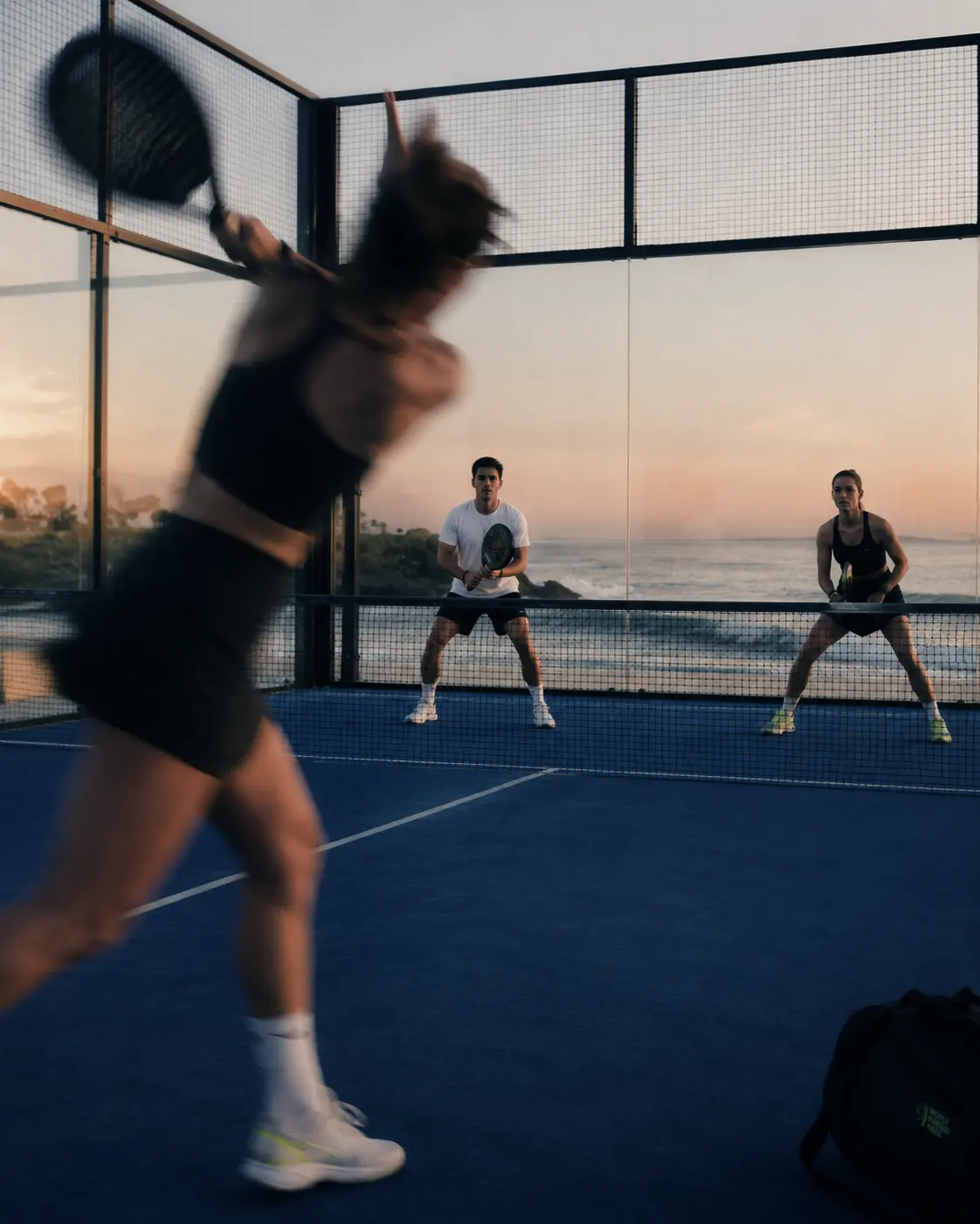 Court No. 02 at dusk, a player mid-smash while two others watch from the far side